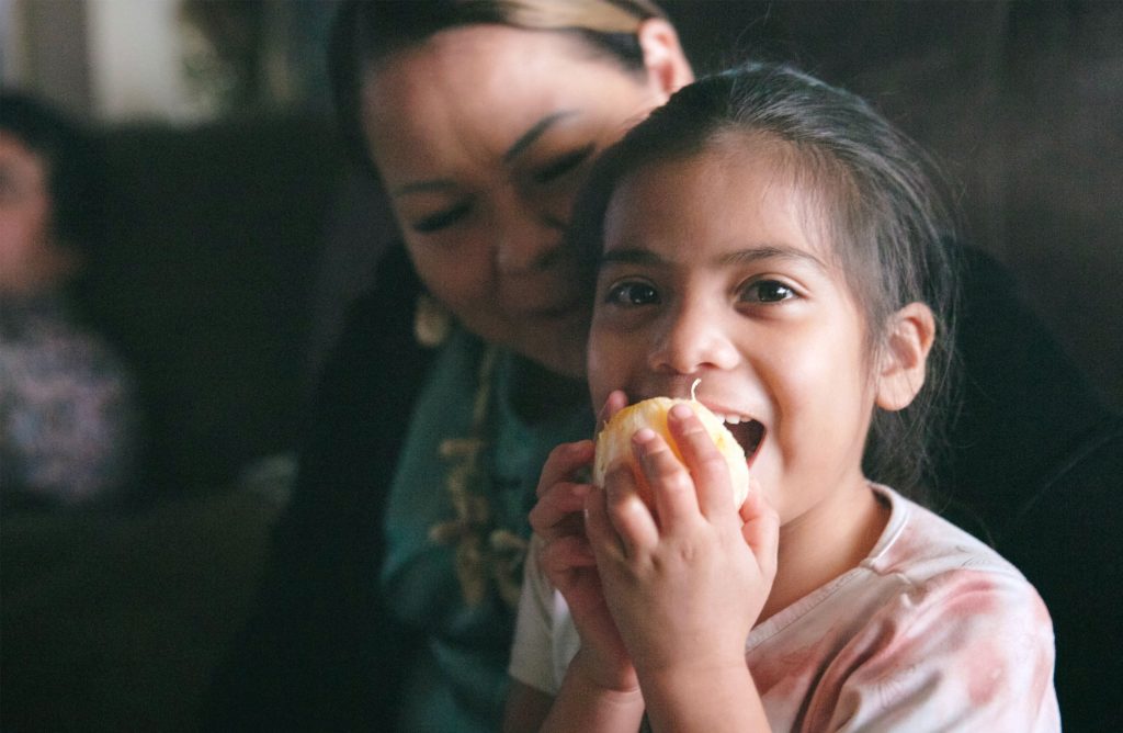 A photo of a girl eating an orange with a woman behind her.