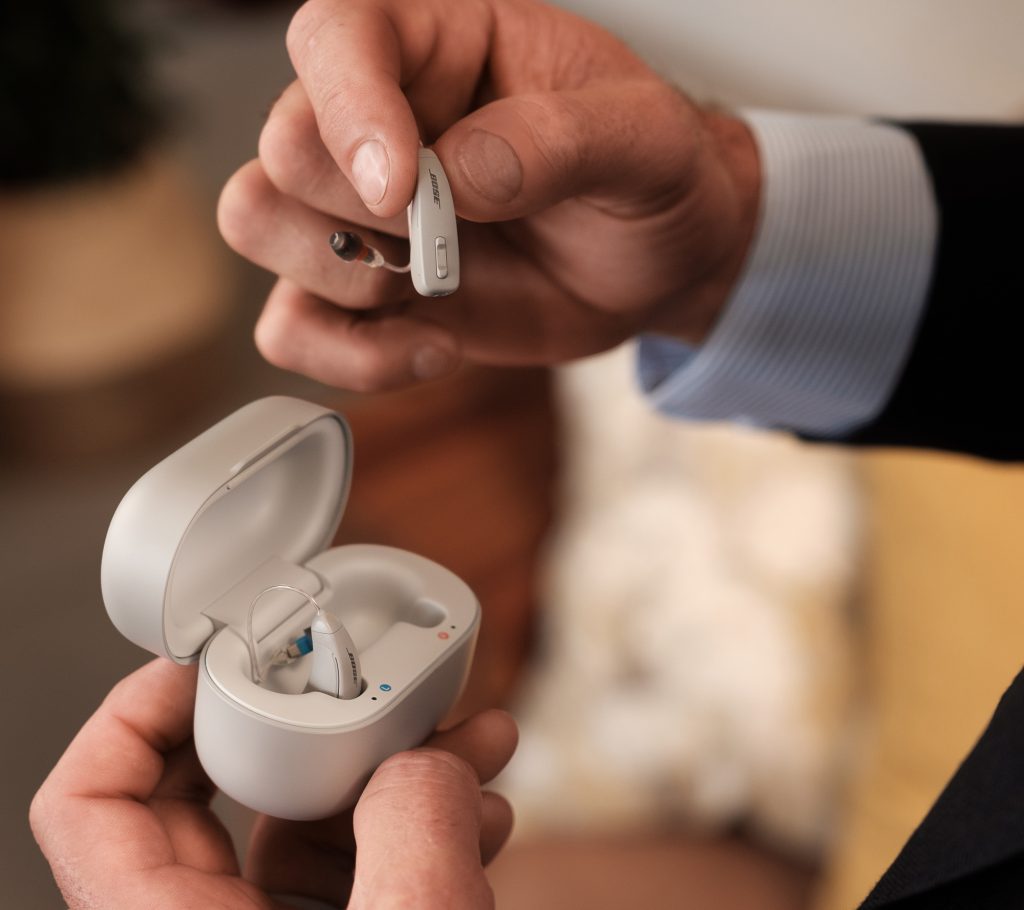 A photo of a man's right hand holding the right Lexie B2 by Bose hearing aid while holding the case in his left hand which contains the left hearing aid,