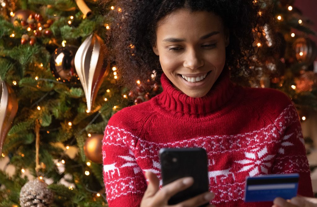 A photo of a woman in a red holiday sweater standing in front of a decorated Christmas Tree holding an iPhone in her right hand and a credit card in her left hand