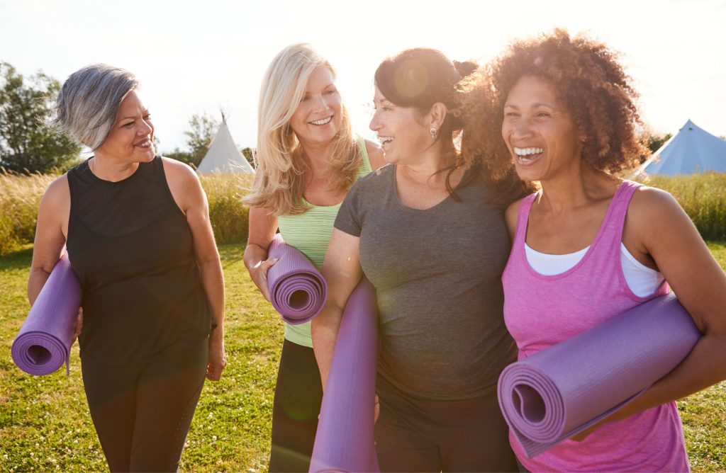 A photo of 4 mature women walking through a field holding yoga mats and laughing