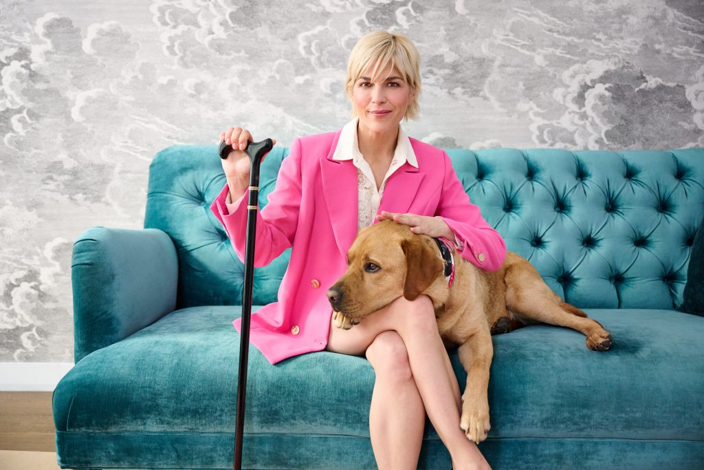 Selma Blair, QVC Brand Ambassador for Accessibility, sits on a plush teal sofa with her dog, Scout. She wears a hot pink suit and uses a black and gold cane.