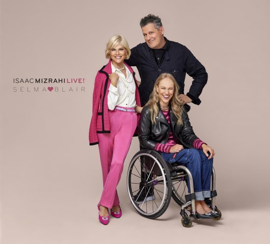 Selma Blair and Isaac Mizrahi, joined by Alex Allen, a QVC model, posing and connected by touch during their photo shoot for the Isaac Mizrahi Live! x Selma Blair QVC collection launch. Selma wears a pink blazer with a white blouse, a pearl necklace, pink pants, and heels. Alex models wheelchair-fit denim jeans, a black moto jacket, and a pink and maroon striped top with black heels. Issac is dressed in dark colors. The background is a color block split screen of neutral and pink.