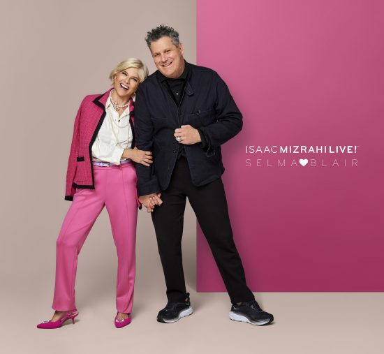 Selma Blair, QVC's Brand Ambassador for Accessibility, stands hand in hand with award-winning fashion designer Isaac Mizrahi, smiling on a photo shoot set. Selma wears a pink blazer with a white blouse, a pearl necklace, pink pants, and heels. Issac is dressed in dark colors. The background is a color block split screen of neutral and pink.